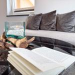 Ein Glas-Tisch mit offenem Buch und Kerze auf Holzstumpf. Hintergrund: Sofa mit Kissen.