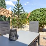 Terrasse mit Tisch und Stühlen, umgeben von Bäumen und Sträuchern unter blauem Himmel.