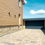 Moderne Villa mit Garagenbereich und gepflastertem Hof unter blauem Himmel.