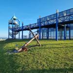 A large, rusty anchor lies on a green lawn in front of a blue building with an observation tower.