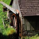 Alte Wassermühle mit Holzrad und Steinmauer an grüner Hanglage.