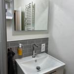 Waschbecken mit Spiegel, Handtuchhalter und Seifenspender an grauer Fliesenspiegelwand.