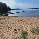 Sandstrand mit Holzsteg zum See. Wald im Hintergrund unter blauem Himmel.