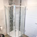 Duschkabine mit Glaswand, Mosaikfliesen und Duschkopf. Weiße Tür und Toilettenpapierhalter.