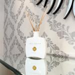 Weißer Duftdiffusor mit Holzstäbchen auf glänzender Oberfläche vor gemustertem Wandbehang.