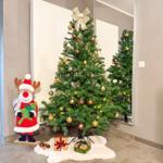 Weihnachtsbaum mit goldenen Kugeln und Schleife, daneben Rentierfigur und Geschenke auf Fellteppich.