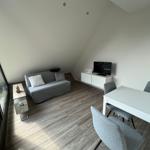 Wohnraum mit Sofa, Esstisch und Fernseher an weißer Wand. Holzdielenboden und große Glasfront.