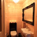 Holzbadezimmer mit Spiegel, Waschbecken und Toilette. Deckenleuchte und Kerzen sorgen für Licht.
