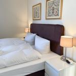 Doppelbett mit weißen Bettwäsche, zwei Nachttische und zwei Lampen. An der Wand hängen zwei gerahmte Bilder.