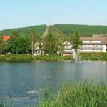 See mit Springbrunnen, umgeben von Grünflächen und Gebäuden. Im Hintergrund bewaldeter Berg mit Aussichtsturm.