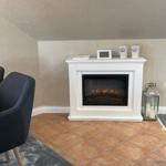 Weißer Kamin mit brennendem Kaminofen, daneben Stehlampe und Sitzgelegenheit.