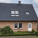 Ein zweistöckiges Haus mit rotem Backstein und dunklem Dach. Vorgarten mit Beeten und Sträuchern.