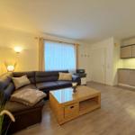 Wohnzimmer mit Sofa, Couchtisch und Küchenbereich. Fenster mit Vorhängen und Bodenbelag.