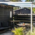 Terrace with seating and table under a canopy. Black wooden wall and plants in the foreground.