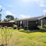A dark wooden house with terrace, garden, and barbecue area under blue sky.