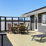 Deck with wooden table, chairs, and lounge chair. Background: house with windows and view of landscape.