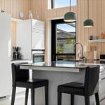 Moderne Küche mit weißen Schränken, schwarzen Barhockern und Holzwänden. Große Fenster mit Blick auf Garten.