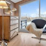 Ein Raum mit Holzwand, Schrank, Stehlampe und Sessel vor großem Fenster mit Aussicht.