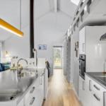 Modern kitchen with countertop, sink, and cooktop. Living area with sofa and windows.