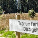 Sign indicates disc golf at Tranum Ferieby. Behind it lies a field with grass and trees.