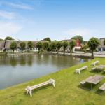 Grüner Rasen mit Picknicktischen und Bänken am See. Dahinter liegen Häuser mit Strohdächern.