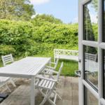 Terrasse mit weißem Tisch und Stühlen, umgeben von grüner Vegetation und einer Bank.