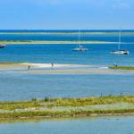 Sailboats und Boots auf ruhigem Wasser, Menschen am Strand und grüne Inseln im Hintergrund.