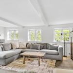 Wohnzimmer mit großem grauem Sofa, Holztisch und Fenstern mit Blick auf Grün.