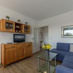 Wohnzimmer mit blauem Sofa, Glas-Tisch und Holzmöbeln. Fernseher und Pflanzen sind sichtbar.