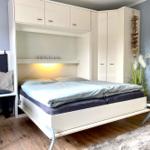 Schlafzimmer mit Bett, Schrank und Sitzgelegenheit. Wandfarbe blau, Holzboden und Teppich.