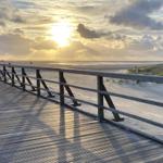 Holzpromenade führt zum Strand bei Sonnenuntergang mit dramatischen Wolken.