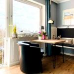 Ein Raum mit Tisch, Sitzbank und Stuhl. Durch Fenster und Tür ist Außenbereich sichtbar.