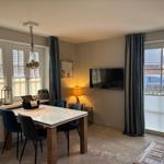Dining area with table, chairs, and TV on wall. Large windows with curtains.