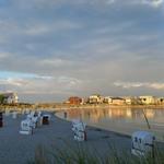 Beach with beach chairs and houses by the water. Sunlight reflects on the water surface.