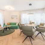 Wohn- und Essbereich mit grünem Sofa, runden Tisch und Stühlen, Holzboden und Fenstern mit Vorhängen.