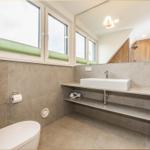 Badezimmer mit Waschtisch, Spiegel, Toilette und Fenstern.