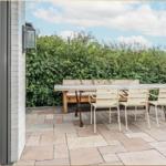 Terrasse mit Holztisch, drei Stühlen und Sitzbank. Hintergrund: grüne Hecke und blauer Himmel.