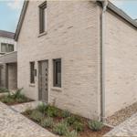 Moderne Villa mit Steinmauer, grauer Tür und gepflastertem Hof. Gartenbereich mit Pflanzen.