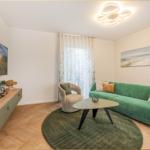 Ein Wohnzimmer mit grünem Sofa, runden Teppich und Wandbild. Fernseher und Sessel sind vorhanden.