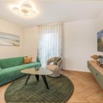 Ein Wohnzimmer mit grünem Sofa, runden Teppich und Wandbild. Fernseher und Couch sichtbar.