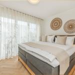 Schlafzimmer mit Doppelbett, Holzfußboden und weißen Wänden. Vorhänge mit Pflanzenmotiv und Wanddekoration.