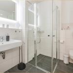 Badezimmer mit Dusche, Waschbecken und Toilette. Weiße Fliesen und graue Bodenfliesen.