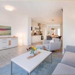 Wohnbereich mit Sofa, Sessel und Esstisch. Küche im Hintergrund. Holzboden und weiße Wände.