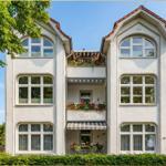 Villa mit drei Etagen, Blumen auf Balkonen und rotem Ziegeldach unter blauem Himmel.