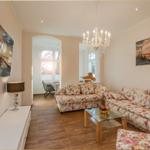 Wohnzimmer mit floralen Sofas, Glas-Tisch und Kronleuchter. Wandbild und Fernseher an der Wand.