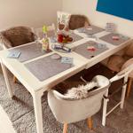 Weißer Tisch mit Stühlen, Platzdeckchen und Obst. Wandbild und Teppich sichtbar.