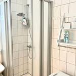 Badezimmer mit Dusche, Waschbecken und Regal. Fliesenwand und Spiegel sichtbar.