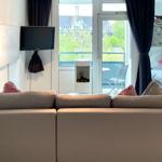 Ein Sofa mit Kissen vor Fenster und Balkon. Fernseher an Wand. Blick auf Haus und Grün.