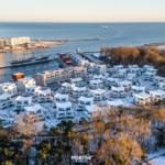 Schneebedeckte Häuser am Hafen mit Blick aufs Meer und einen Segler.