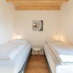 Zwei Betten mit weißen Laken, Holzboden und Deckenbalken. Zwischen den Betten steht ein kleiner Tisch.